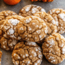 Soft, golden-brown pumpkin crinkle cookies with dramatic white powdered sugar cracks, arranged on a cooling rack with warm autumn lighting