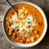 A bowl of creamy orange-colored chicken pumpkin chili topped with shredded cheese and green onions, served with crusty bread on a wooden table with fall decorations in the background.