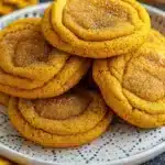 Stack of golden-brown pumpkin spice cookies with cinnamon sugar coating on rustic wooden board, surrounded by autumn leaves and whole spices