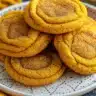 Stack of golden-brown pumpkin spice cookies with cinnamon sugar coating on rustic wooden board, surrounded by autumn leaves and whole spices