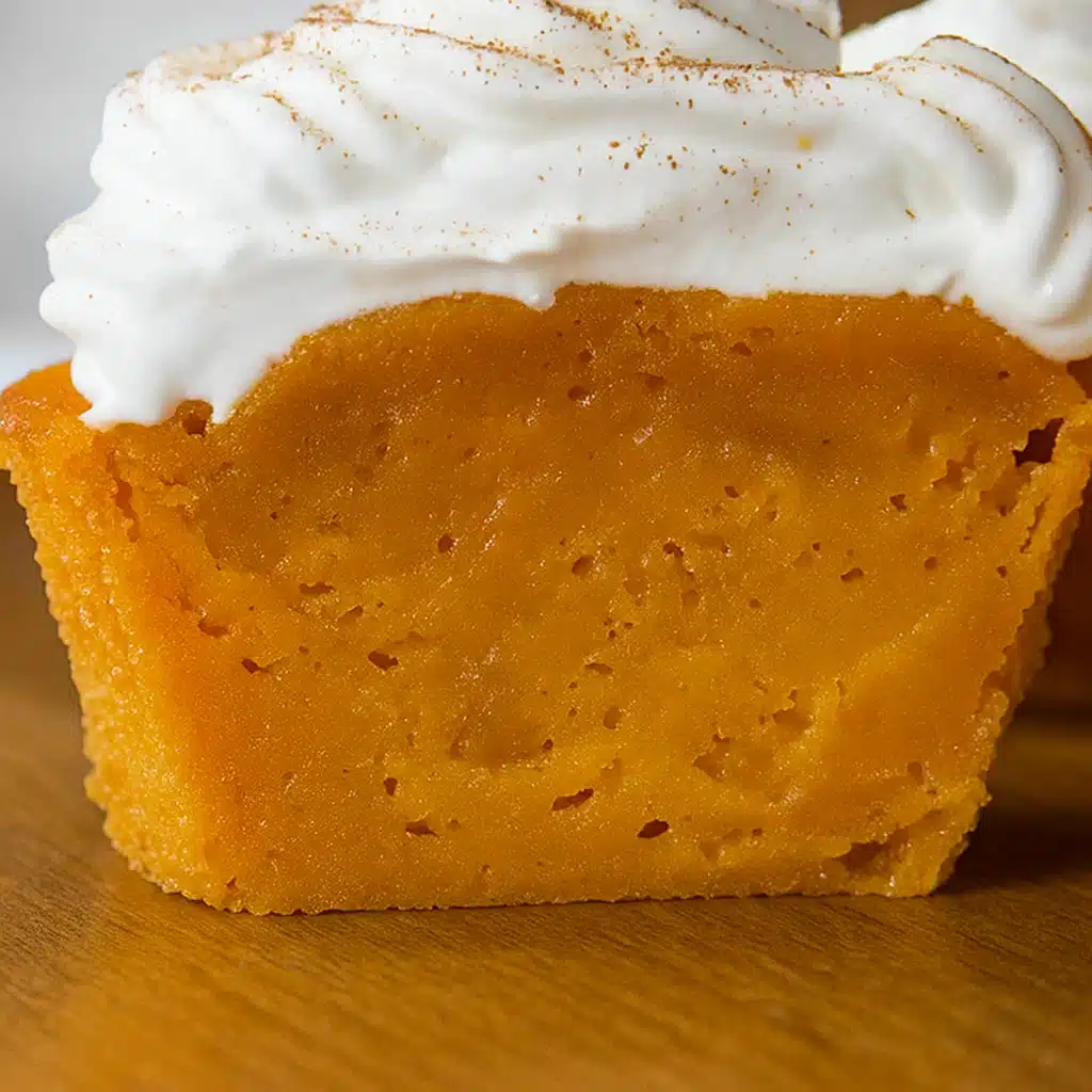 brown impossible pumpkin pie cupcakes with white whipped cream dollops and cinnamon dust, sitting on a wooden cutting board with autumn leaves scattered around.