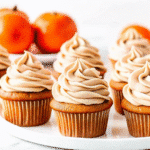 Perfectly frosted pumpkin spice cupcakes with cream cheese frosting, dusted with pumpkin pie spice, arranged on a rustic wooden board with fall leaves